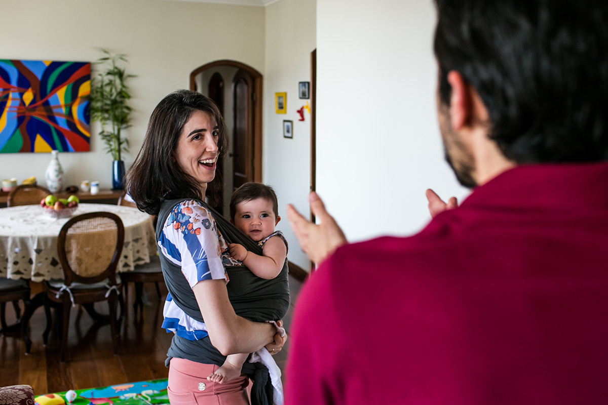 pai brincando com a filha que está no colo da mãe com o wrap sling, em foto de Adriana Costa
