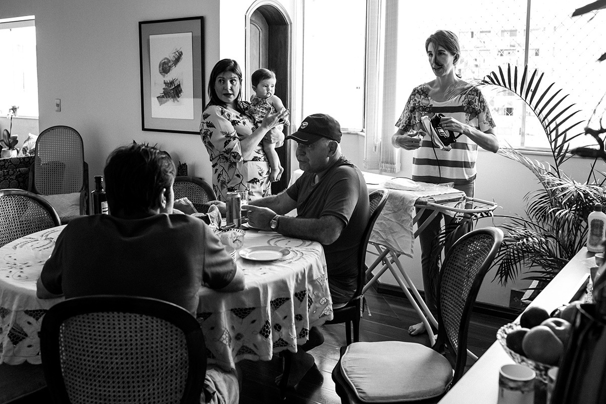 mãe preparando a mesa de passar para engomar as lembrancinhas do batizado da sua filha, enquanto a família se prepara para almoçar