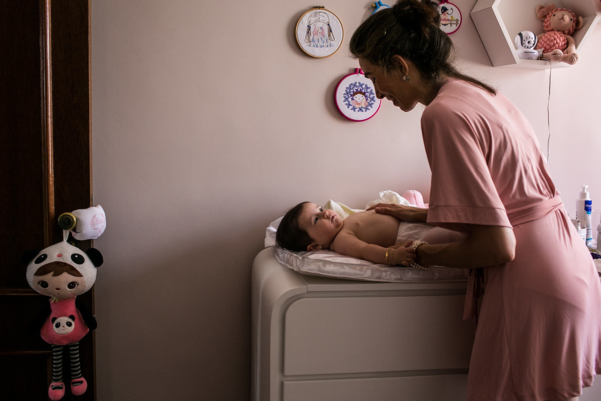 mãe começando a vestir sua filha para a cerimônia de batismo da sua filha, por Adriana Costa