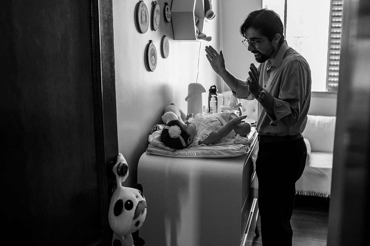 Pai brincando com a filha enquanto a mãe se arruma para o batizado da filha, em foto de Adriana Costa