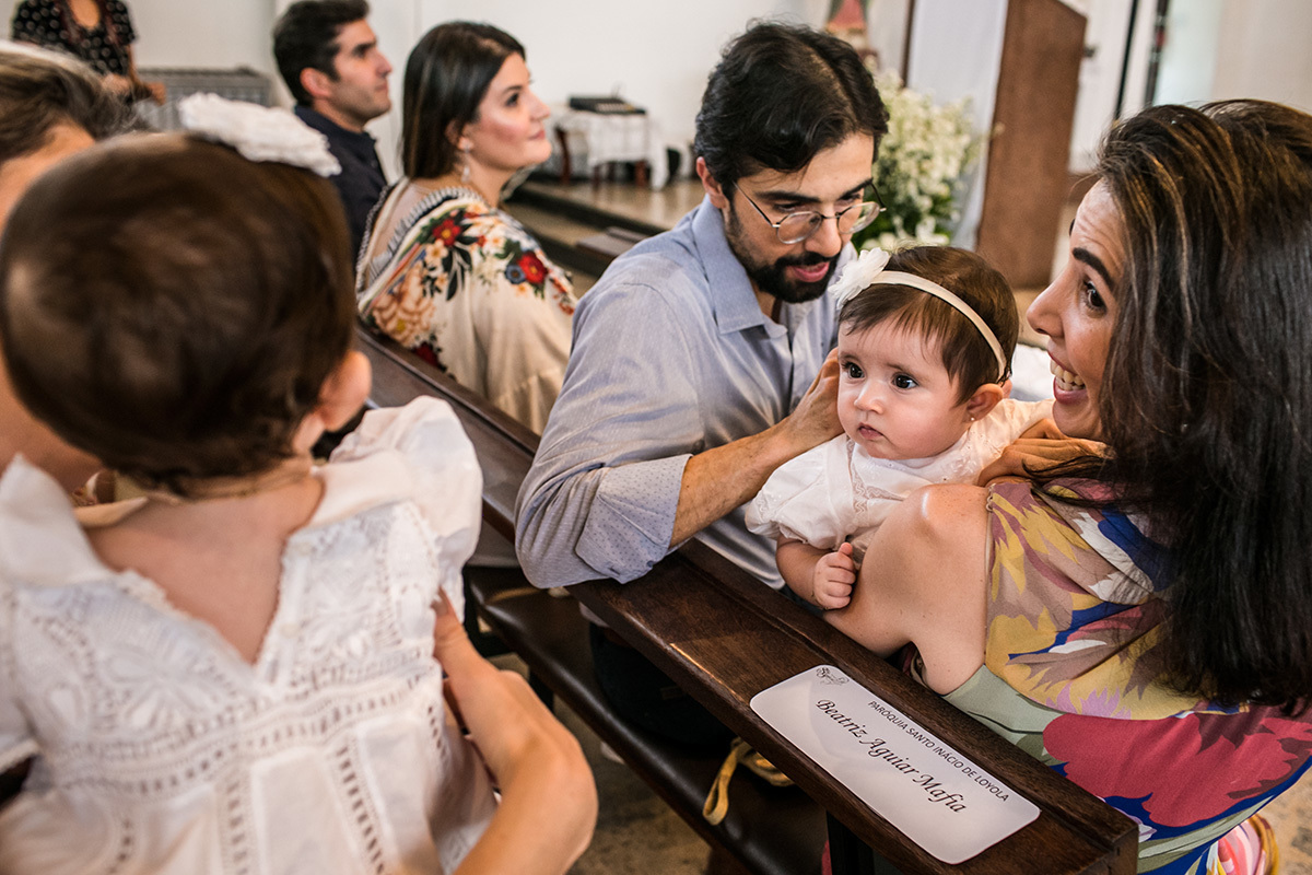 pai abotoa a bata de batismo da filha ou mandrião enquanto ela brinca com o bebê que está no banco de trás, por Adriana Costa