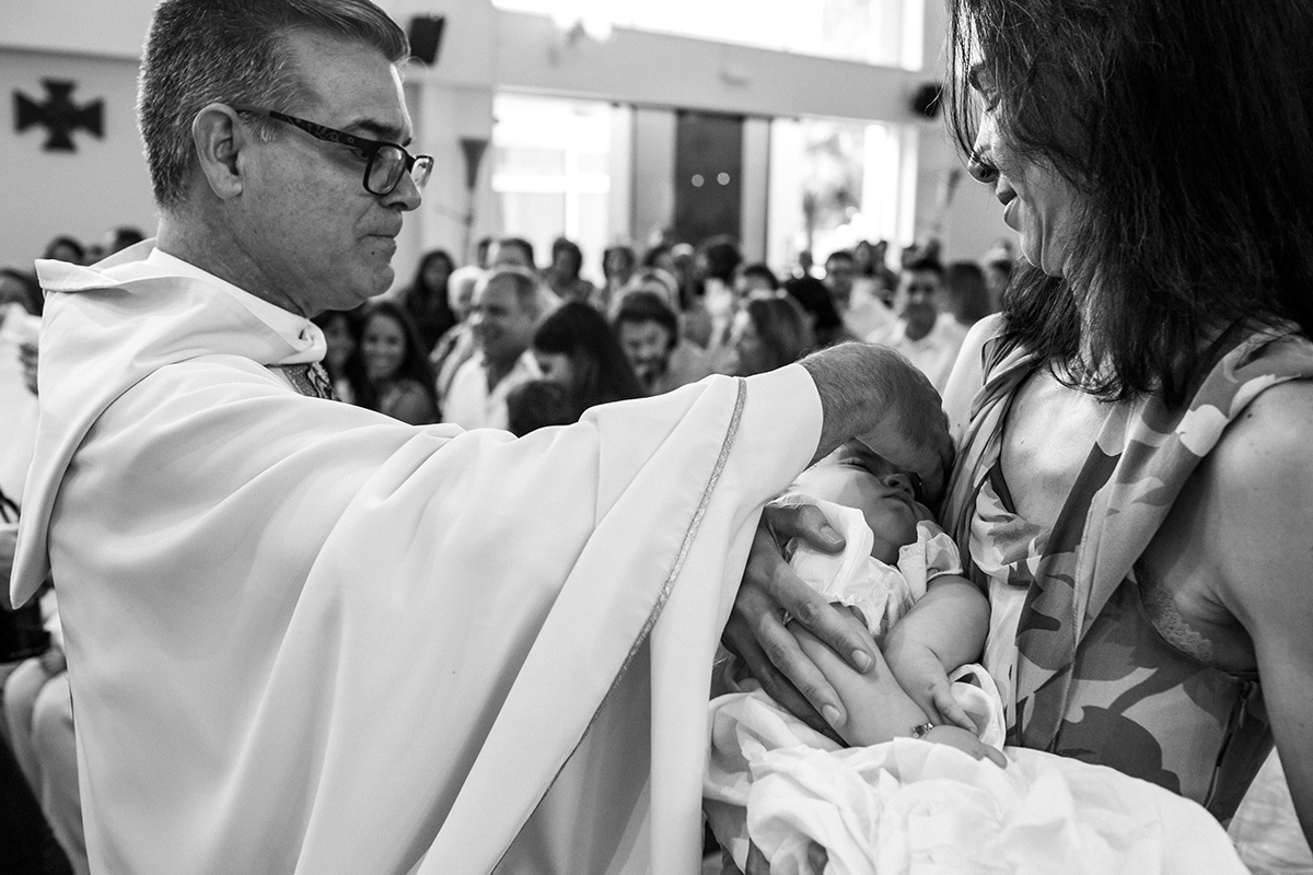 Adriana Costa registra padre passando água benta na cabecinha da bebê que dorme durante sua cerimônia de batismo