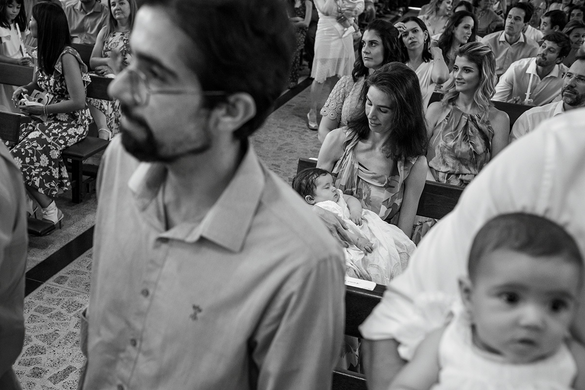 pai espera ser abençoado pelo padre enquanto a mãe espera com sua bebê que está dormindo no bando da igreja, por Adriana Costa