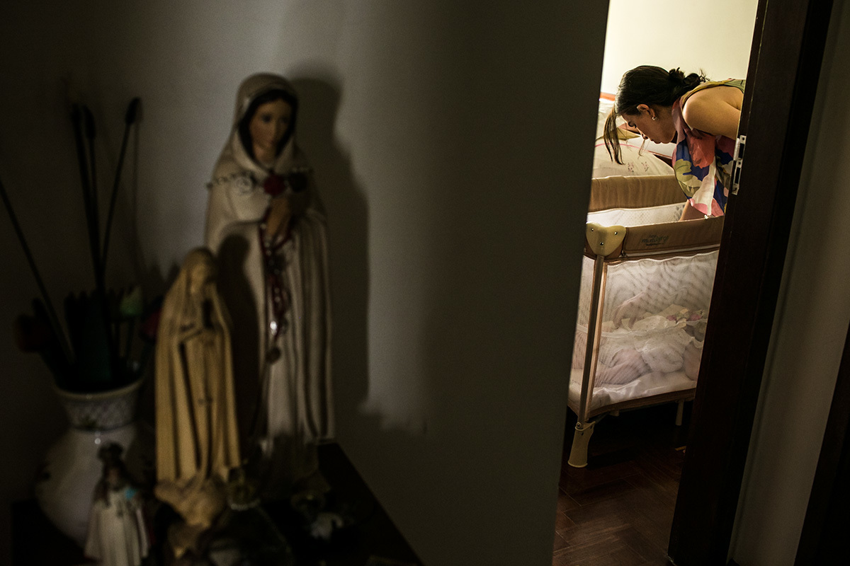 mãe colocando sua bebê no berço desmontável que fica na casa da avó próximo a uma mesa com imagens de santa, após a festa de batizado, por Adriana Costa