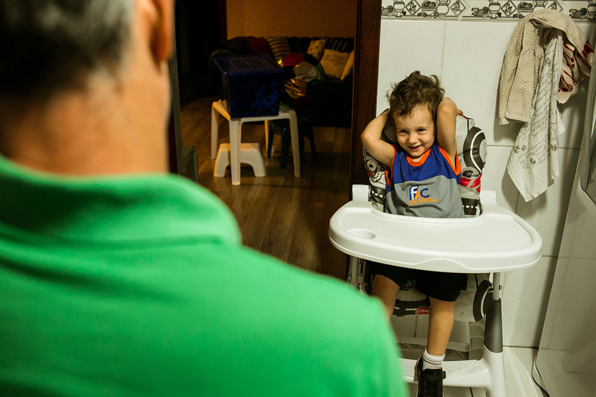 menino na cadeirinha de comer rindo para o pai na hora do jantar, por Adriana Costa