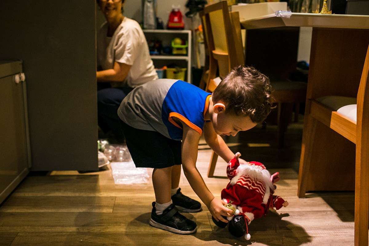 menino brinca com um Papai Noel, enquanto sua mãe pega enfeites de Natal no armário