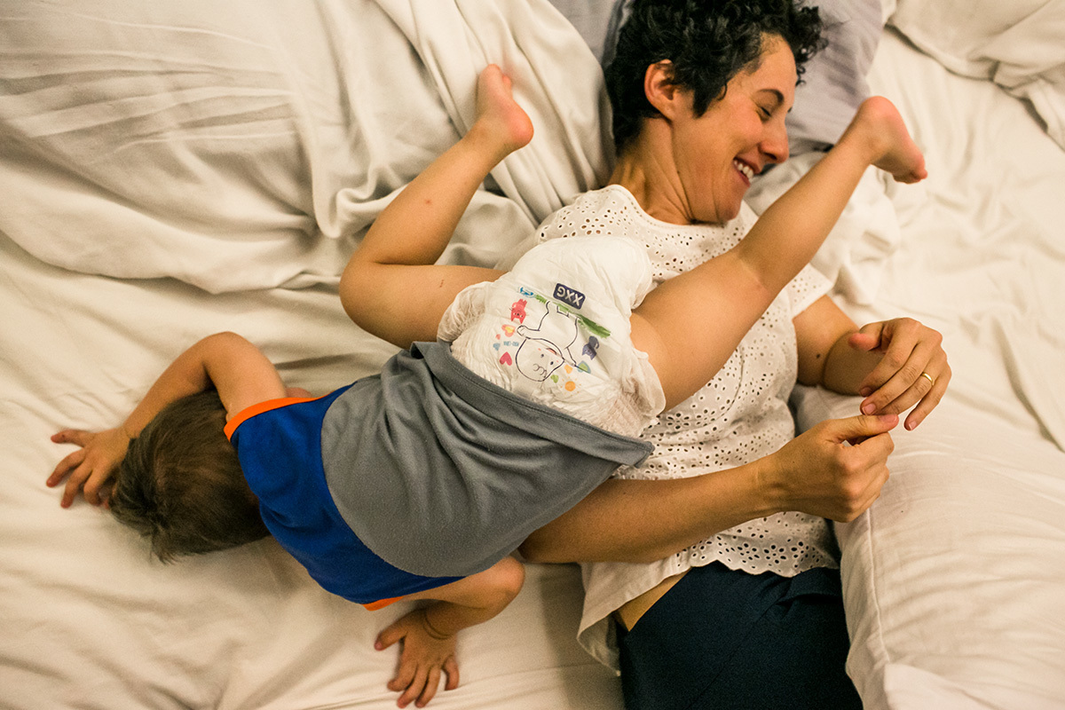 filho pula por cima da mãe enquanto brincam na cama, em foto de Adriana Costa