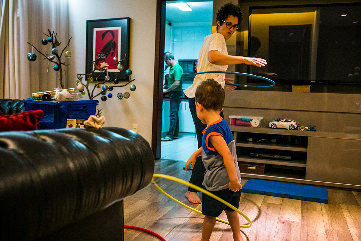mãe e menino de 2 anos brincam de bambolê em casa enquanto o pai prepara o lanche, por Adriana Costa