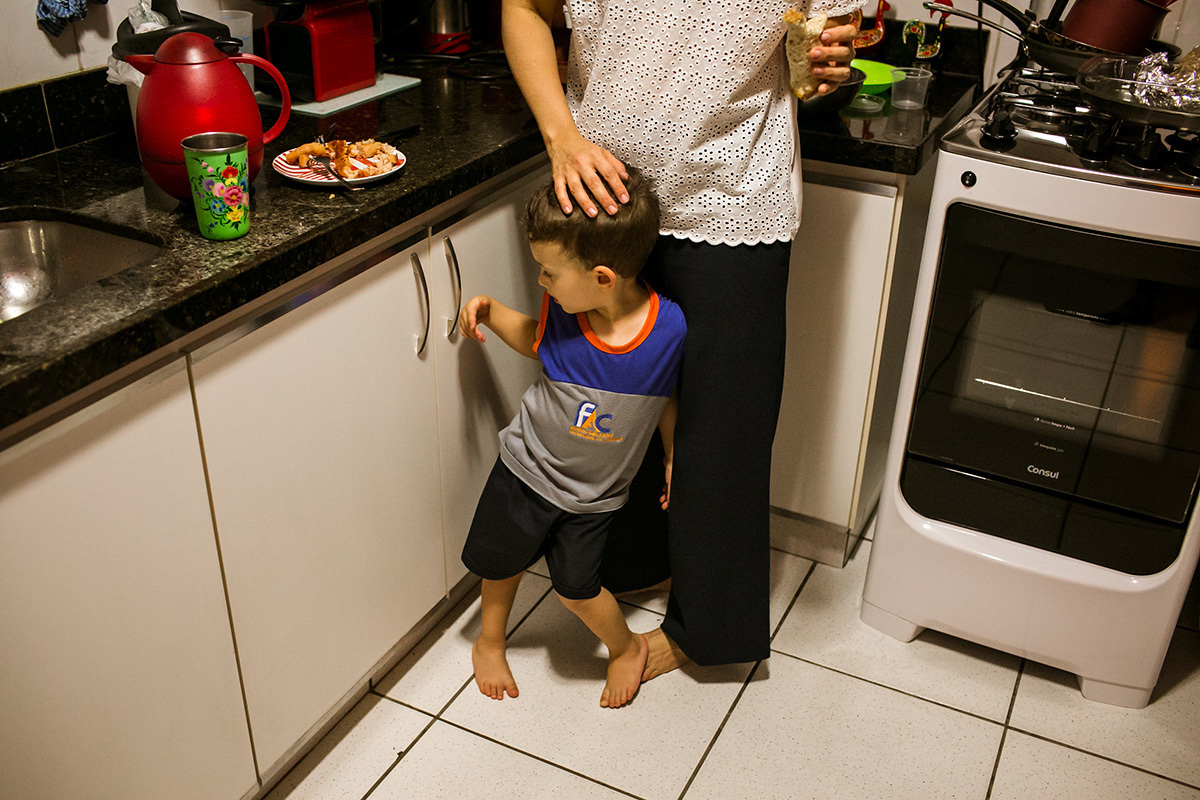 menino fica em cima do pé da mãe enquanto ela come um lanche na cozinha