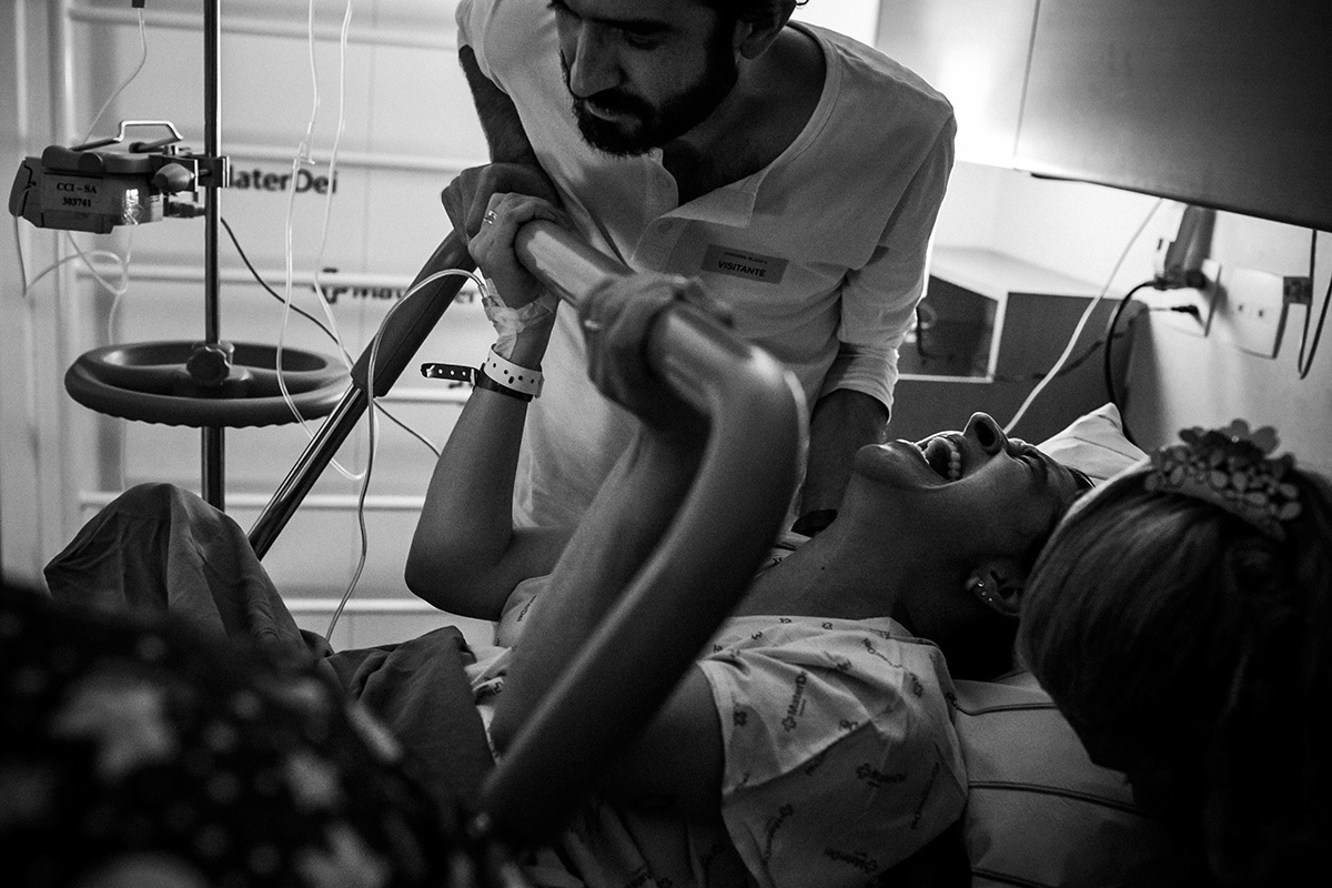 marido observa se seu bebê está nascendo enquanto sua esposa faz força para ele sair, por Adriana Costa