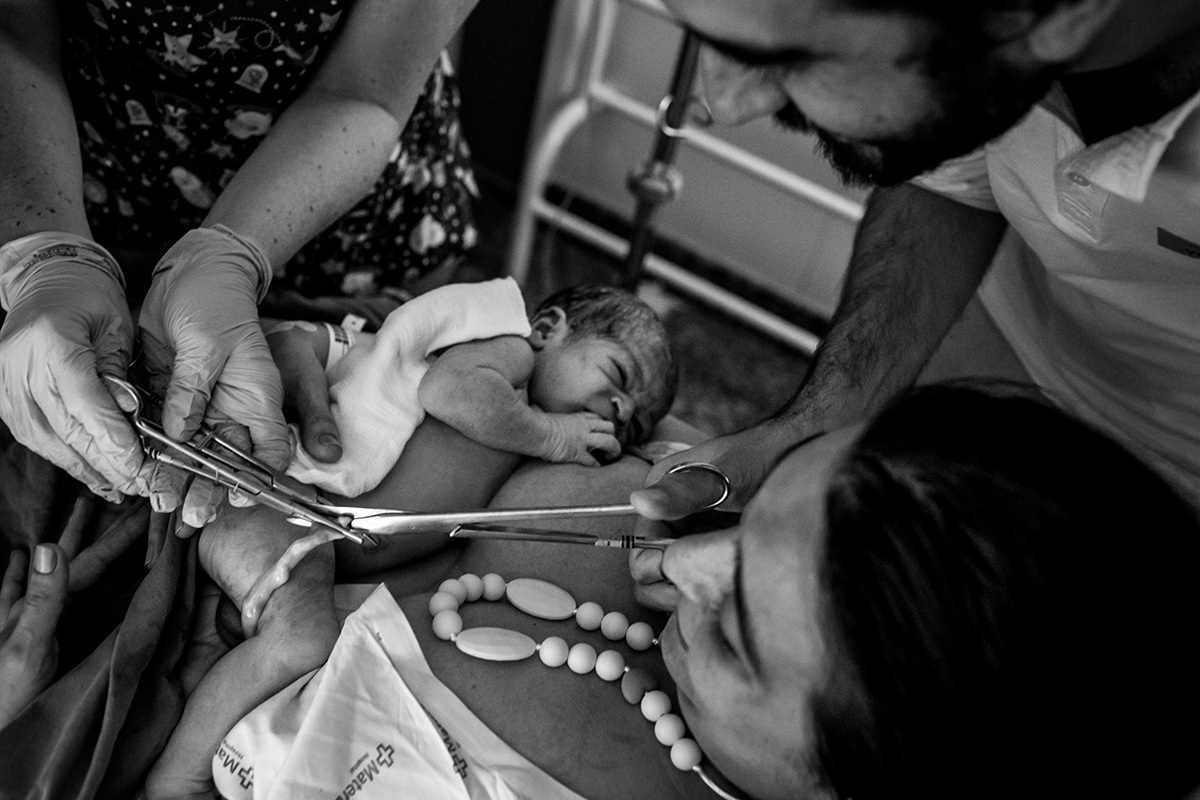 pai corta o cordão umbilical do filho que está no colo da mãe