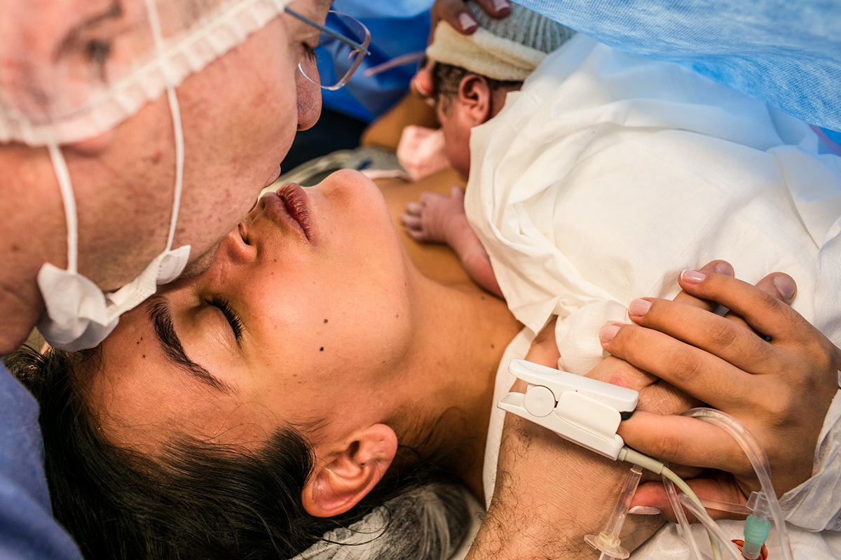 Foto de Adriana Costa dos pais se beijando emocionados com sua filha nos braços após cesárea