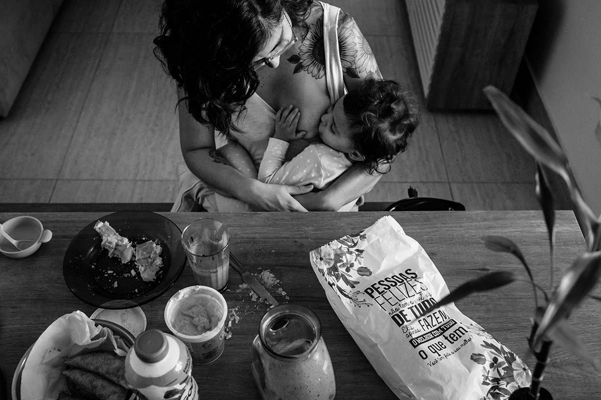 criança amamenta durante o café da manhã no colo da mãe, em foto de Adriana Costa