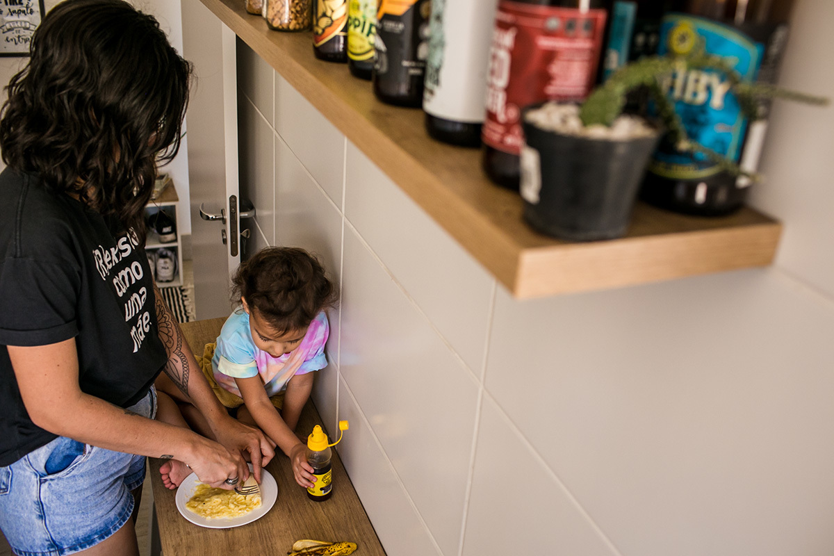 foto de Adriana Costa da menina assentada na bancada da cozinha para ajudar a mãe a prepara a banana para ela comer