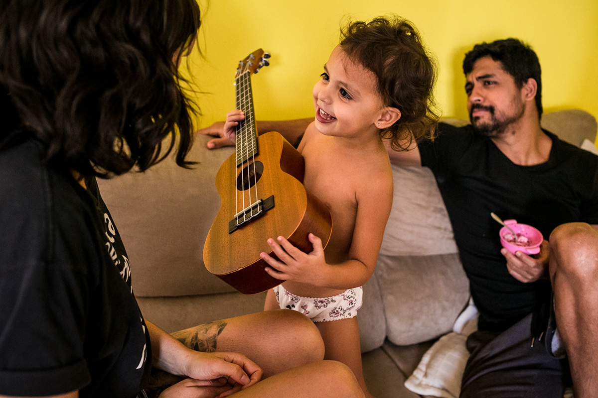 filha pede para tocar  ukulele para a mãe sob o olhar atento e divertido do pai, por Adriana Costa