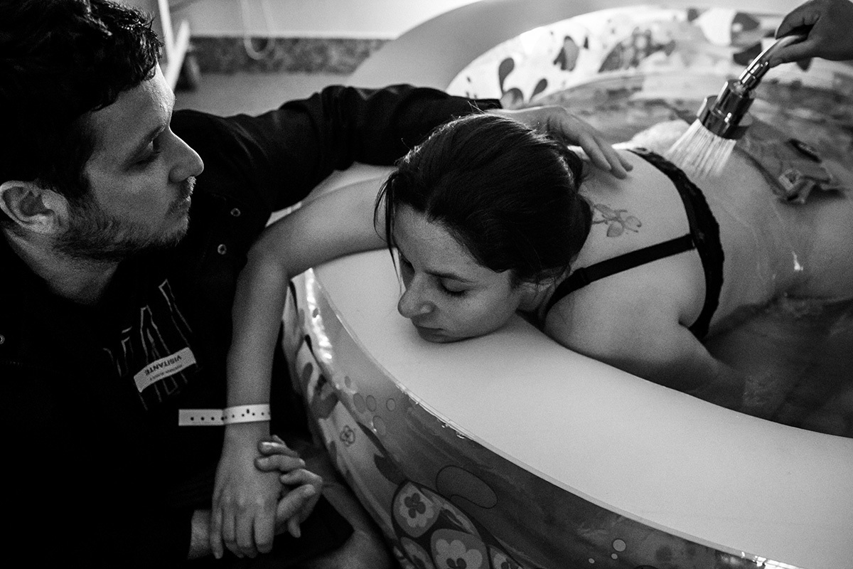 marido fazendo carinho na mulher que se encontra na banheira para relaxar durante o trabalho de parto, em foto de Adriana Costa