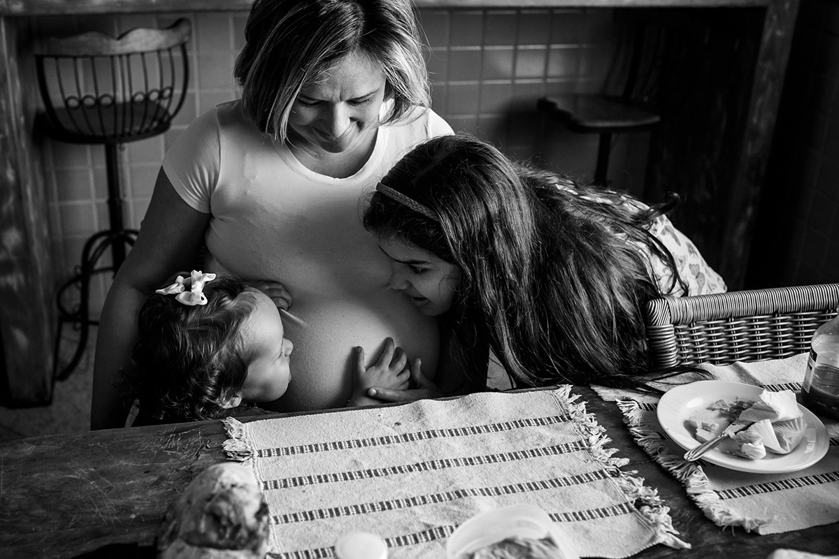 mãe grávida no café da manhã brincando com suas duas outras filhas em foto de Adriana Costa