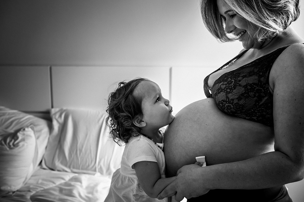 criança abraçada a barriga de sua mãe gravida da terceira filha em foto de Adriana Costa