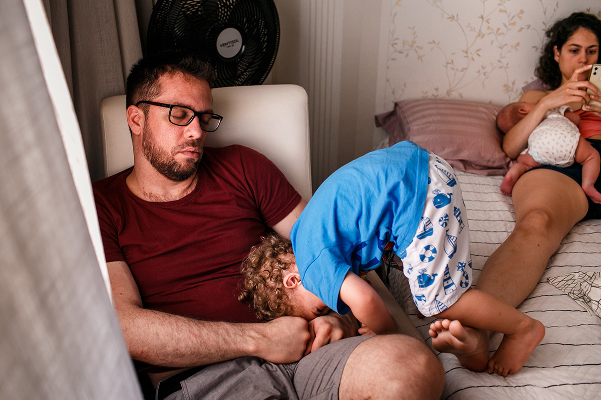 menino tenta acordar o pai enquanto sua mãe amamenta seu irmão recém-nascido em foto de Adriana Costa