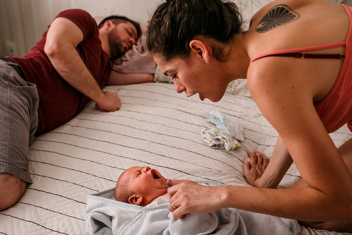 bebê recém-nascido bocejando enquanto sua mãe o enxuga do banho e o seu pai dorme 