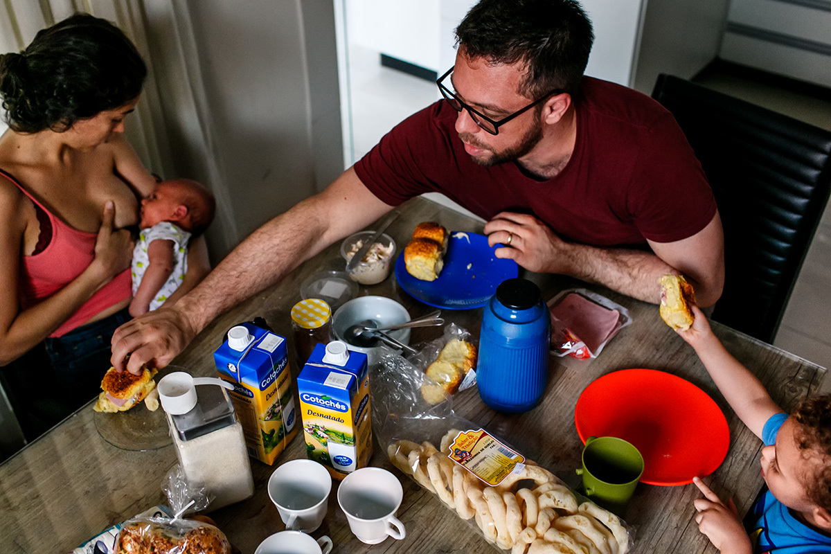 homem ajuda esposa que está amamentando o seu filho mais velho na hora do lanche em foto de Adriana Costa
