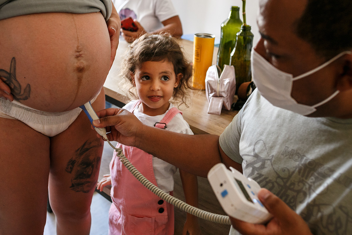 Foto de Adriana Costa da criança observando o enfermeiro da equipe Bom Parto examinar sua mãe durante o trabalho de parto domiciliar