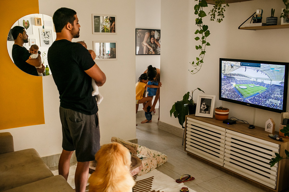 pai cuida do bebê assistindo futebol enquanto a mãe troca a roupa da filha mais velha