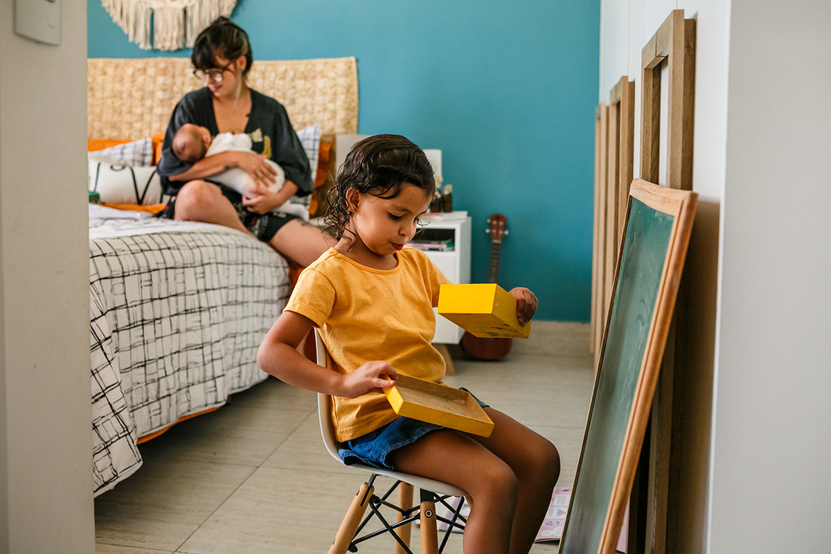 criança brinca ao lado da mãe que está amamentando seu bebê em foto de Adriana Costa