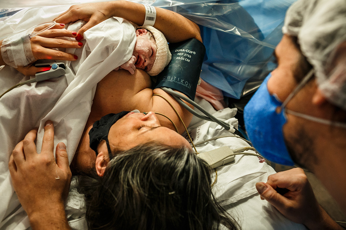 mãe permanece com sua filha nos braços enquanto o obstetra finaliza a cesárea em foto de Adriana Costa