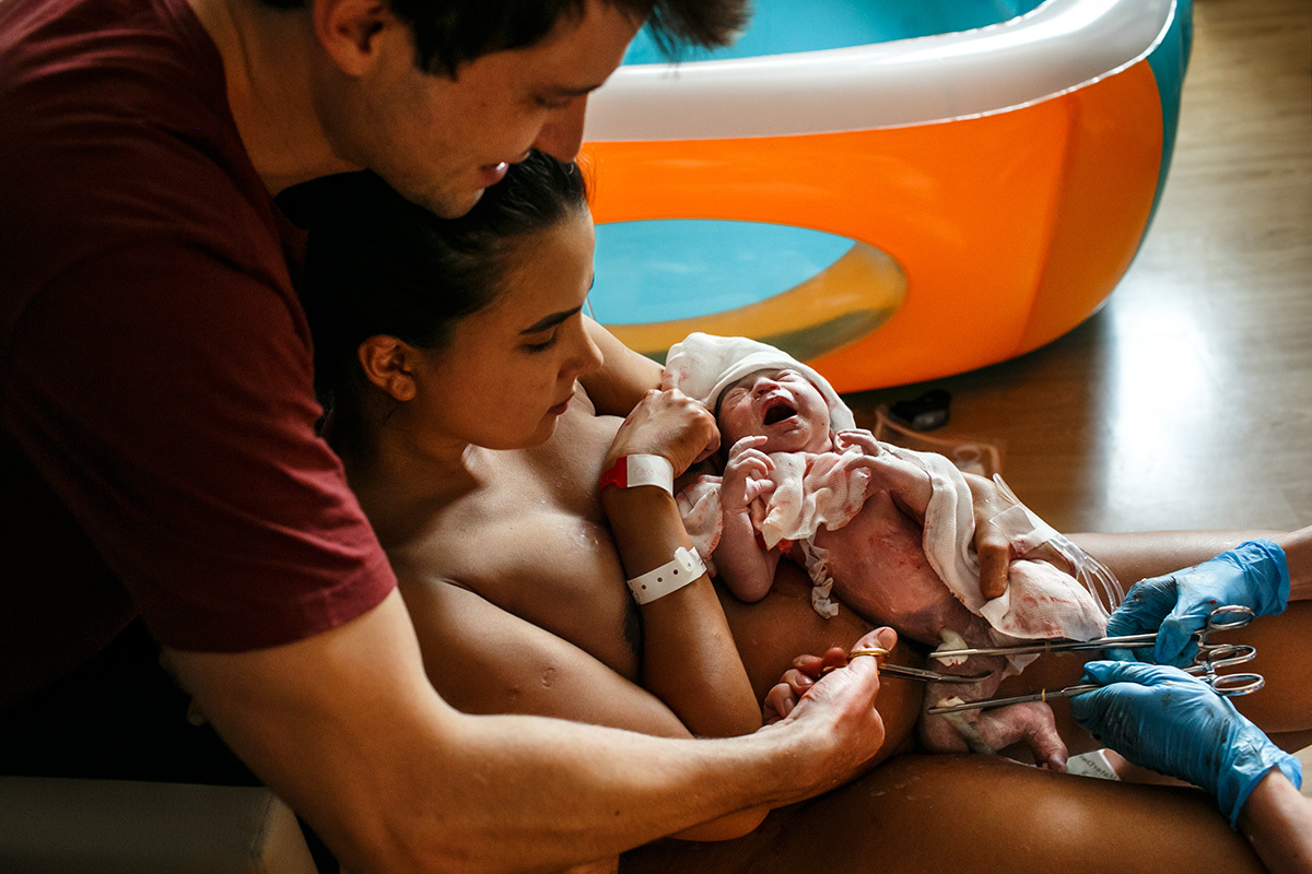pai corta o cordão umbilical da sua bebê com a mãe ainda assentada na banqueta do parto