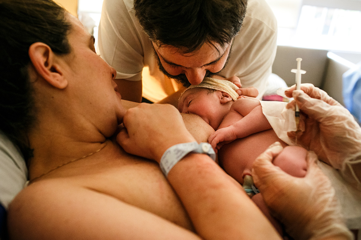 foto de Adriana Costa do pai dando beijo em seu bebê que acabou de nascer enquanto ele amamenta e recebe a vitamina K injetável 