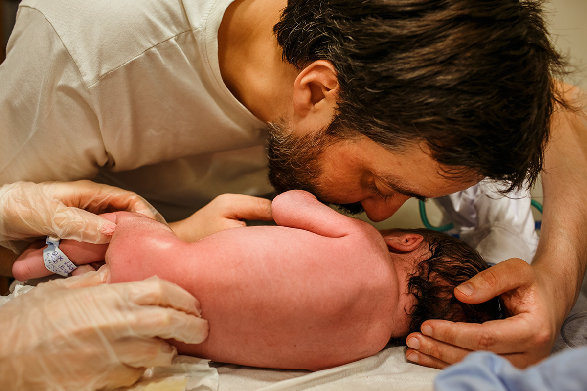 foto de Adriana Costa do pai falando no ouvido de seu bebê após o nascimento enquanto ele recebe seu primeiro exame