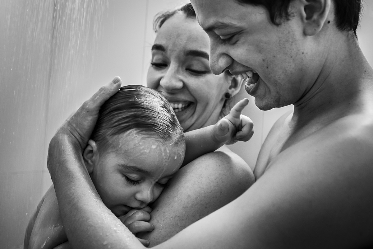 bebê brinca com os pais e ao mesmo tempo cochila durante o banho de chuveiro em foto de Adriana Costa