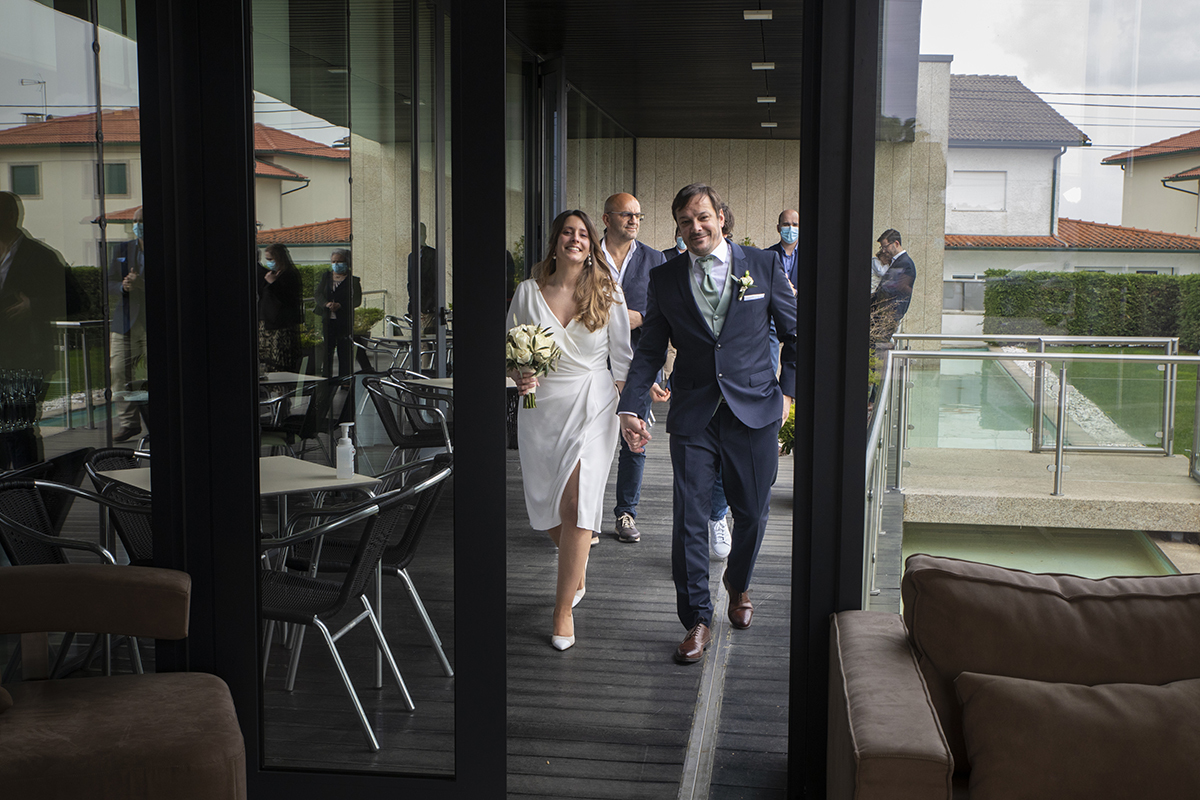 Por uma porta aberta, os noivos caminham descontraídos em direção à câmara. Rita segura o bouquet, e os sorrisos são leves e naturais.