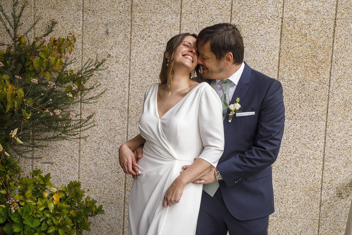 Encostados à parede de pedra, rodeados por vegetação, Nuno abraça Rita pela cintura. Ela encosta a cabeça na dele e sorri, enquanto ele a acompanha no gesto.