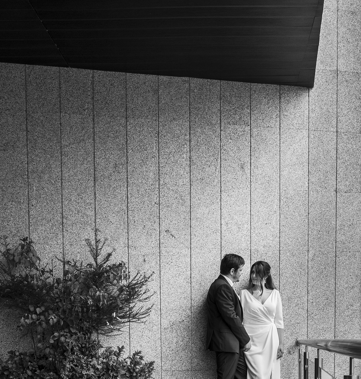 A preto e branco, uma parede de pedra moderna com riscos verticais cria um fundo sofisticado. Ao lado, uma planta verde traz contraste e, no canto direito, Nuno e Rita encostam-se à parede, olhando-se com ternura.