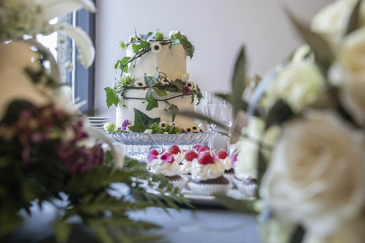 O bolo de casamento surge ao fundo, enquanto, em primeiro plano, arranjos de flores desfocados criam um efeito encantador. No centro da composição, os cupcakes complementam o cenário. Restaurante santa luzia