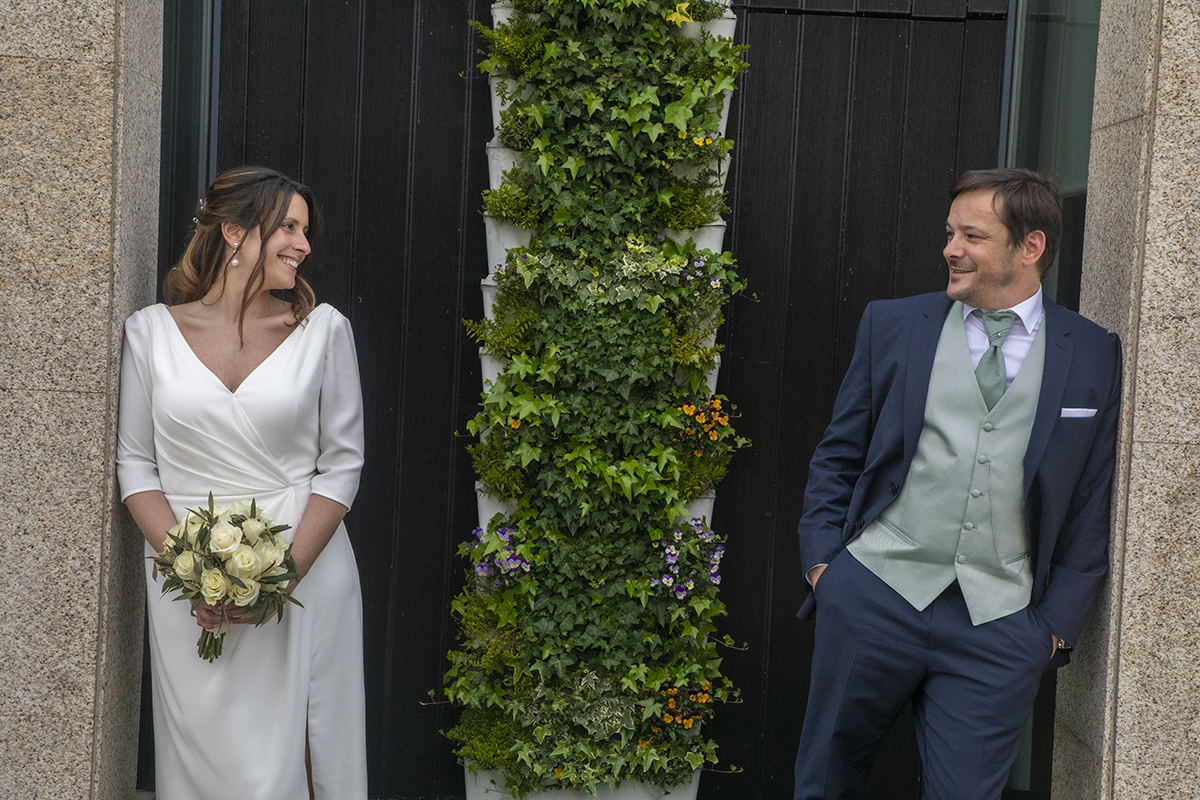 Em frente a uma porta verde decorada com plantas, os noivos encostam-se a pilares opostos, trocando olhares felizes. Rita segura o bouquet delicadamente.