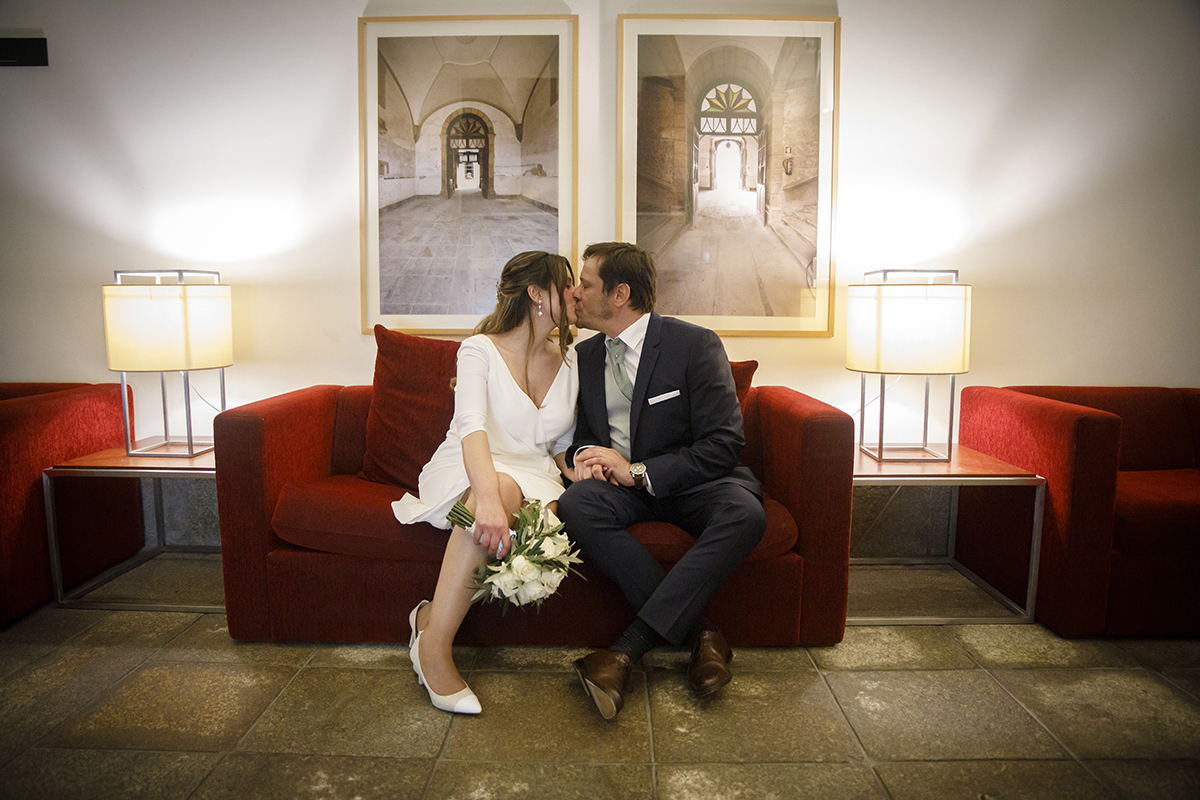 Sentados num sofá vermelho, Nuno e Rita trocam um beijo apaixonado, imersos na elegância do espaço. Pousada de Viseu