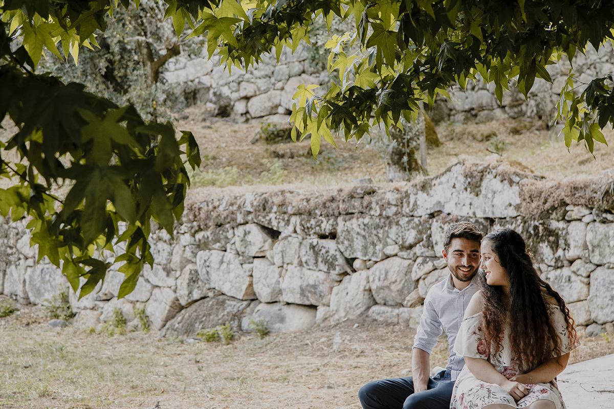 Sentados, Juliana de costas para Miguel mas virando o rosto, e ele sorrindo com admiração, enquanto o muro de pedra ao fundo testemunha este olhar cheio de carinho.