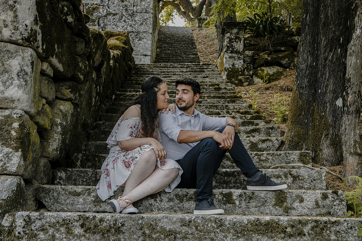 Na escada, Juliana pousa o queixo no ombro de Miguel; olhares trocados entre sorrisos serenos que refletem uma cumplicidade silenciosa e um amor tranquilo.