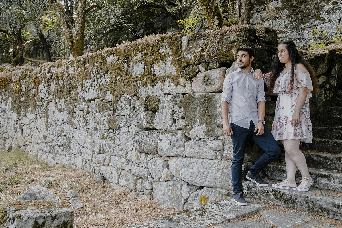 Encostados ao muro de pedra junto à escada, Juliana repousa o braço no ombro de Miguel, enquanto ambos contemplam o horizonte num momento de reflexão profunda.