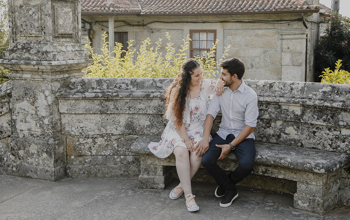 Sentados num banco antigo de pedra, com Juliana de pernas cruzadas e Miguel tocando-lhe o joelho, o amor deles manifesta-se num toque delicado e cheio de significado.