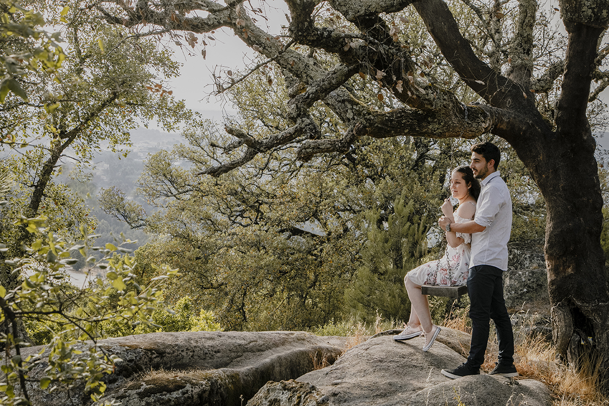 Ao pé de uma árvore com baloiço, Juliana senta-se enquanto Miguel segura as correntes; ambos olham para a frente, serenos e felizes, rodeados pela natureza.