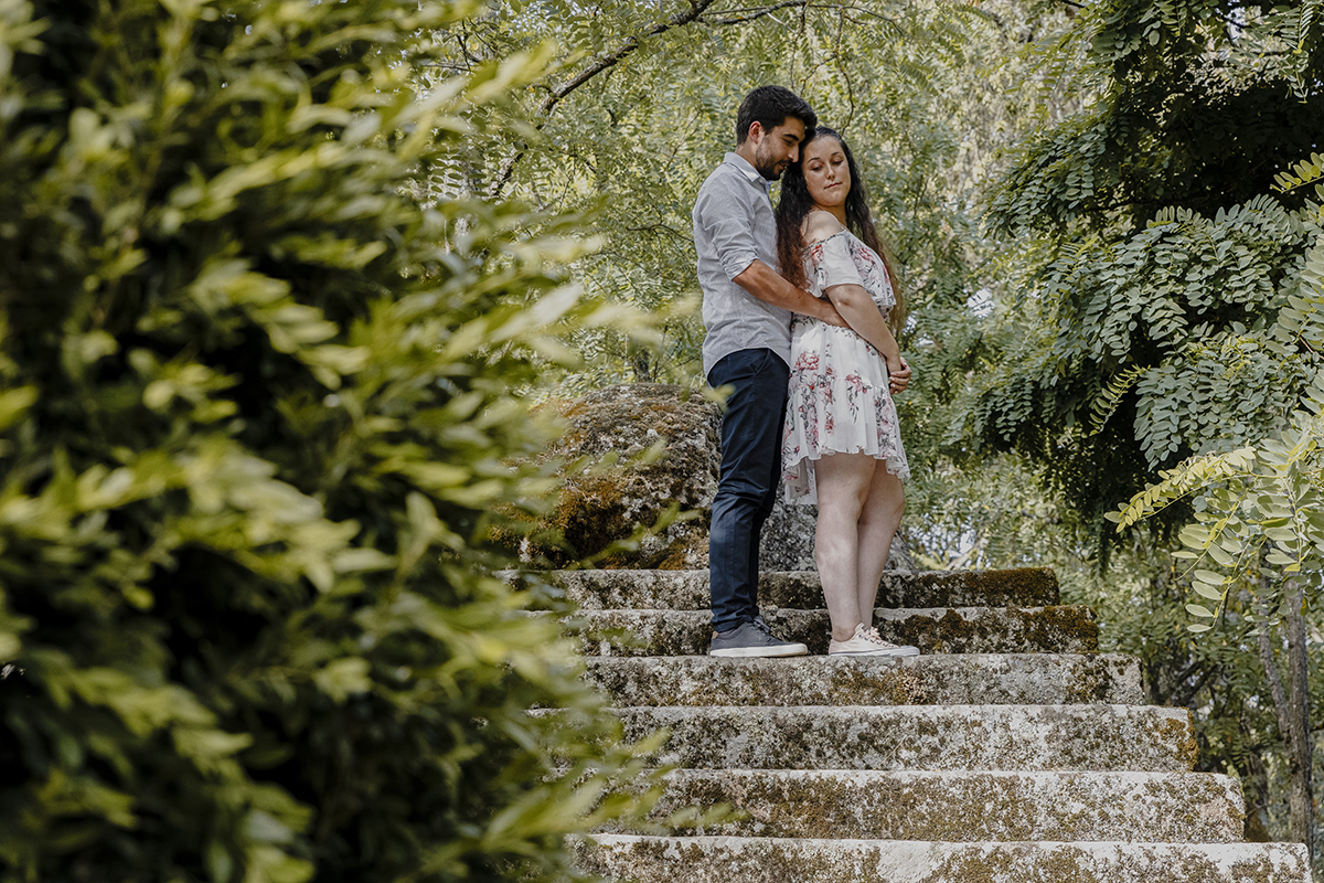 No lado esquerdo, os arbustos verdes desfocados emolduram Miguel e Juliana nas escadas, num abraço íntimo, com as cabeças suavemente encostadas, mostrando um momento de ternura e cumplicidade.
