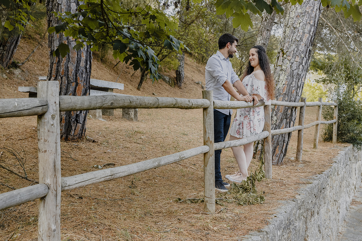 Numa cena mais ampla, o corrimão, as mesas e as árvores são cenário para os sorrisos trocados entre Juliana e Miguel, num momento de pura conexão e alegria partilhada.