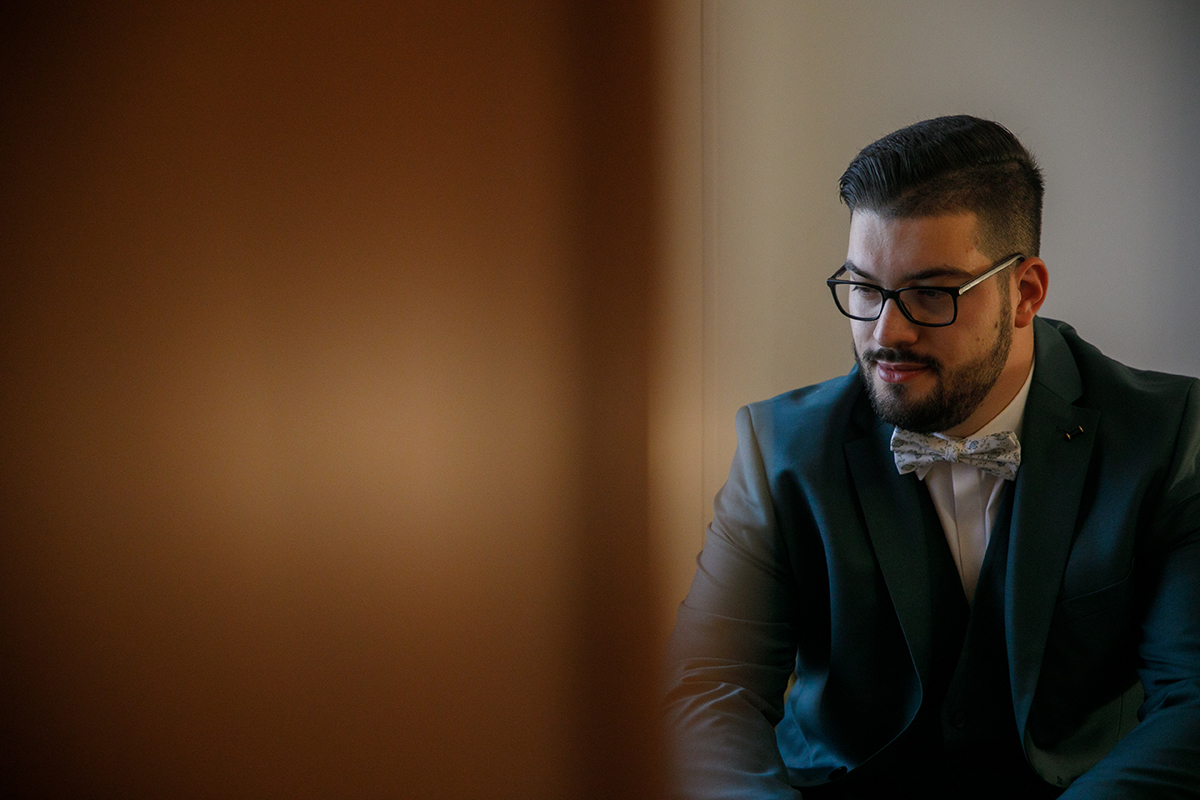 Sentado à direita da imagem, Tiago olha para baixo com um leve sorriso no rosto, quase a sonhar acordado, enquanto do lado esquerdo uma porta de madeira desfocada cria um contraste acolhedor.