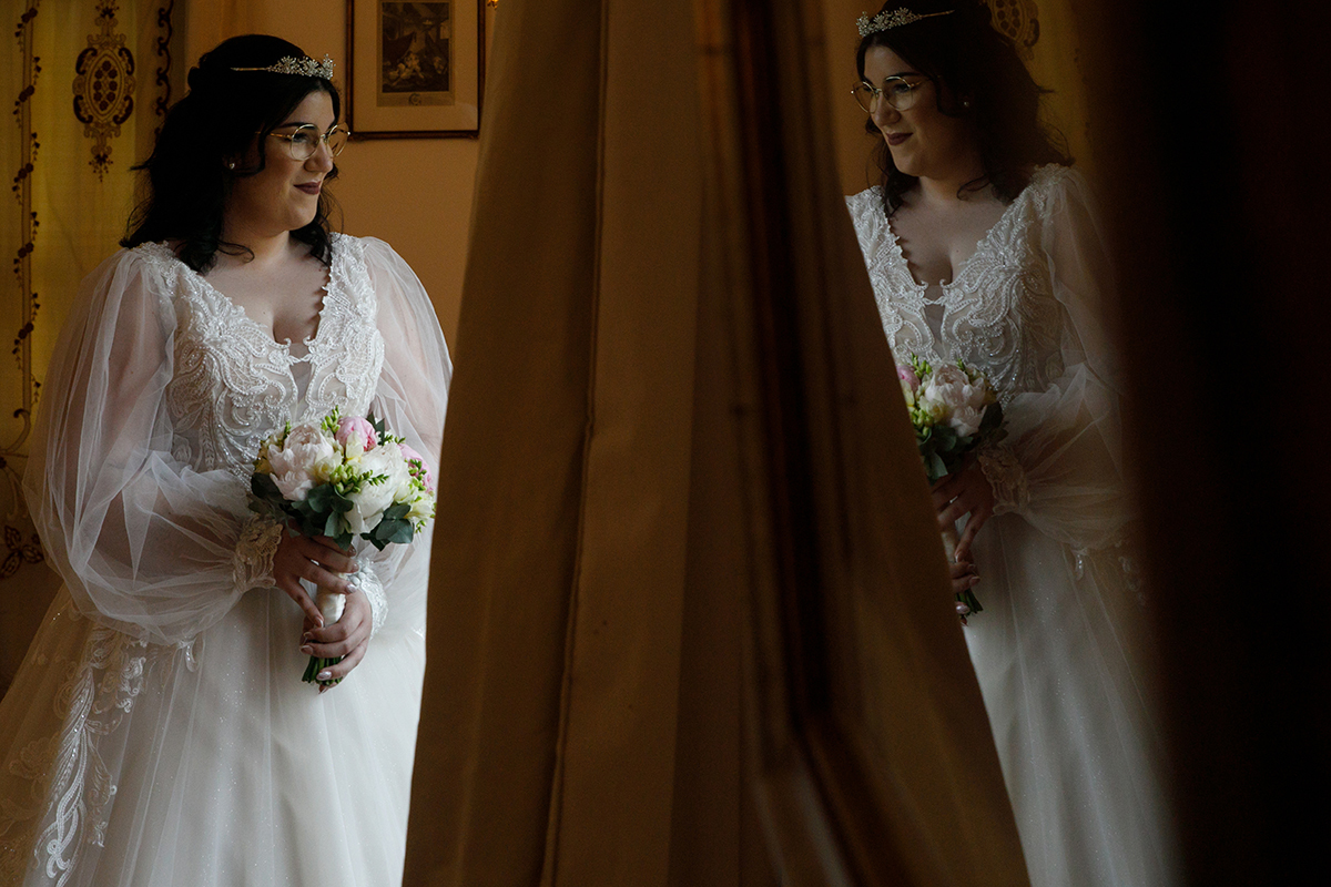 Carolina surge junto à janela segurando o bouquet, o olhar fixo na luz que entra, enquanto o reflexo da sua figura depois da cortina cria um duplo encanto, uma imagem cheia de graça e mistério.