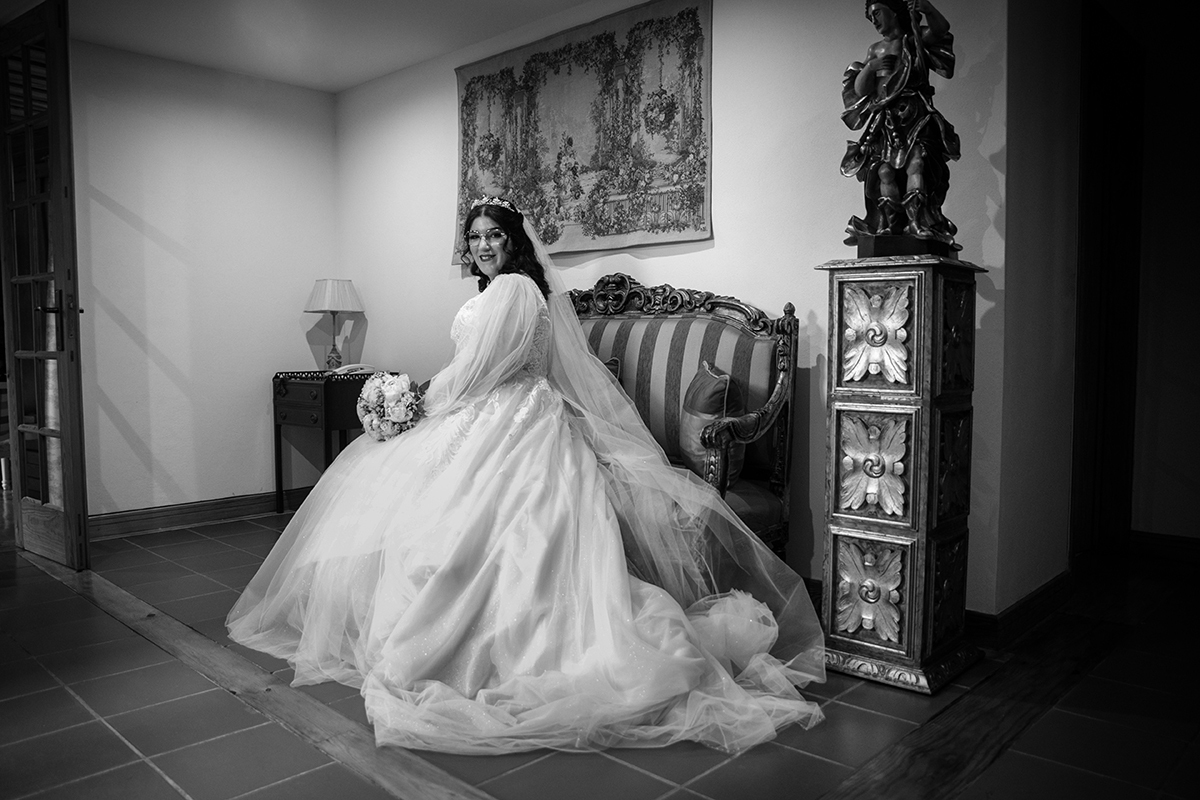 Aqui vemos a Carolina sentada num sofá antigo de tons suaves, a olhar para a câmara com um sorriso sereno enquanto segura delicadamente o seu bouquet repousando sobre as pernas, um momento de calma e beleza pura.