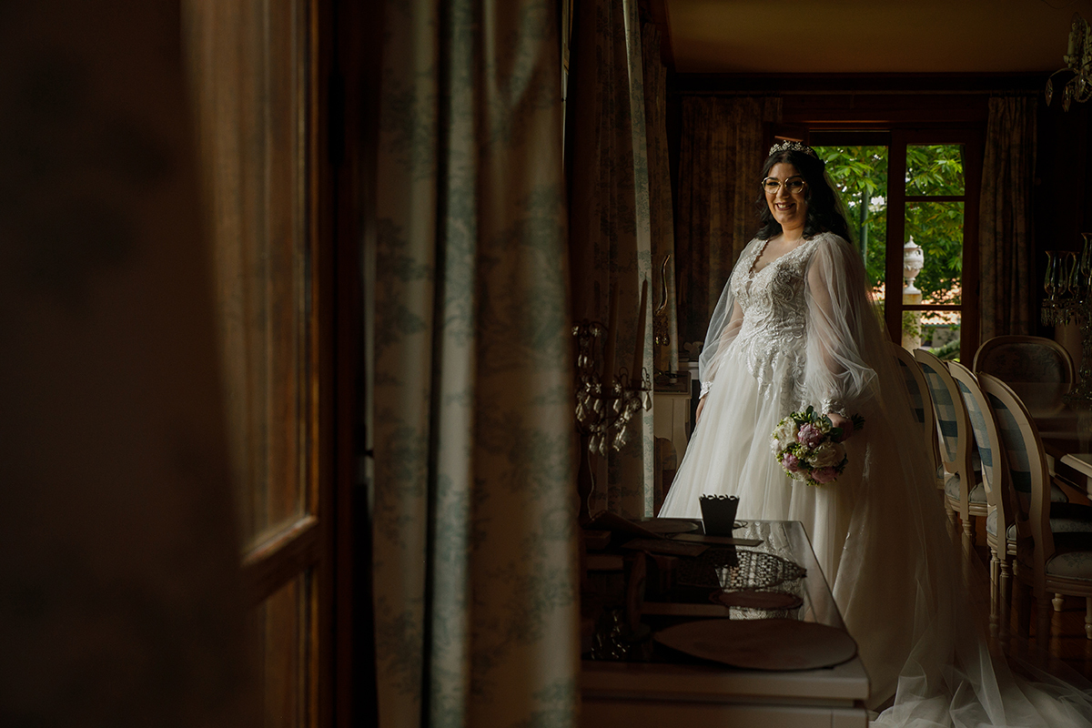 Na sala de jantar, Carolina sorri ligeiramente de lado para a câmara, envolvida pela luz que invade o espaço e pelo brilho do seu vestido deslumbrante, enquanto a mesa e as cadeiras desenham um cenário de elegância discreta.